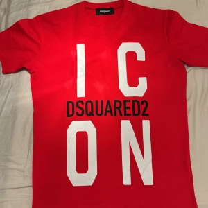 Röd t-shirt från DSQUARED2 - Säljer en snygg röd t-shirt från DSQUARED2 med texten 'ICON' i vitt och märkesnamnet i svart. Perfekt för en casual look eller att sticka ut på festen. T-shirten är i bra skick och har en normal passform. Kortärmad och gjord i mjukt material för bästa komfort. Knappt använd och helt som ny, självklart äkta och köpt från cettire för 1400 fast på rea. Paket från cettire finns kvar.