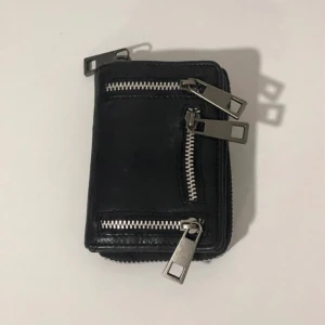 Svart plånbok med dragkedjor - Säljer en snygg svart plånbok i äkta skinn med flera dragkedjor och tryckknappar. Perfekt för att hålla ordning på kort och kontanter. Den är i bra skick och har en stilren design som passar alla tillfällen.