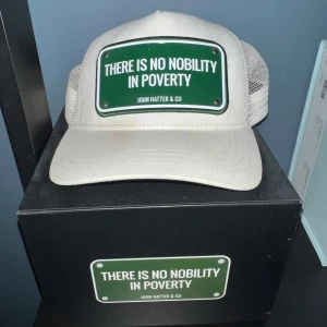 Vit keps från John Hatter & Co - Snygg vit keps från John Hatter & Co med ett grönt märke framtill där det står 'There is no nobility in poverty'. Kepsen har mesh på sidorna och justerbar passform. Hör av er vid minsta lilla! Kan gå med o sänka pris vid snabb affär! Det är plåt inte gummi!