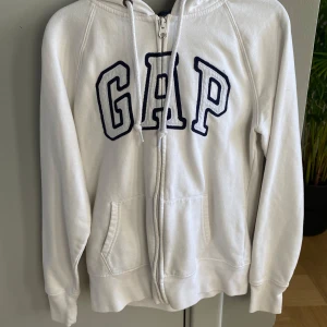 Vit hoodie från GAP - Säljer en supermjuk och bekväm vit hoodie från GAP i storlek XS. Den har en dragkedja framtill och fickor på sidorna. Perfekt för kyliga dagar eller mysiga hemmakvällar. GAP-loggan är broderad i mörkblått på bröstet. Passar till allt och är ett måste i garderoben! 🤍