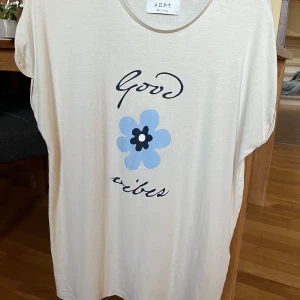 Beige t-shirt med tryck från ADPT - Säljer en superfin beige t-shirt från ADPT med texten 'Good Vibes' och en blå blomma på framsidan. Den är i mjukt material och har en avslappnad passform. Perfekt för en chill dag eller sommarhäng! 🌼