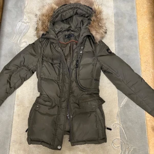 Olivgrön dunjacka från Hollies - Säljer en olivgrön dunjacka från Hollies i storlek 36. Jackan har en mysig pälskantad huva och långa ärmar. Perfekt för kalla vinterdagar med sin varma vaddering och stilrena design. Den har praktiska fickor och en dragkedja framtill. Passar både till vardags och lite finare tillfällen.