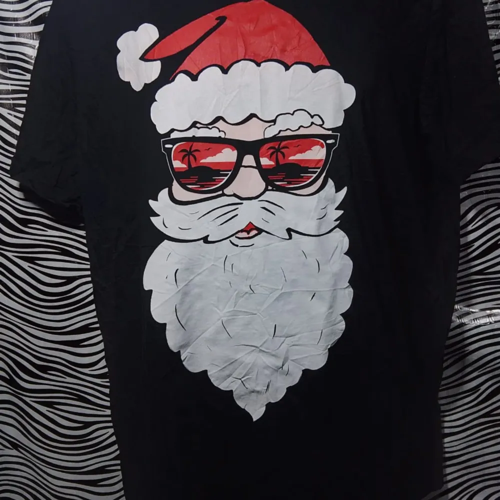 Cool svart t-shirt med ett stort tryck av en jultomte som bär solglasögon med en strandreflektion. Perfekt för att sprida julstämning med en twist! T-shirten är kortärmad och gjord i mjukt material. Perfekt för både jul och sommarvibbar.. T-shirts.