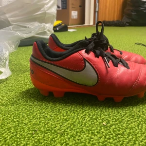 Röda Nike Tiempo fotbollsskor - Säljer ett par röda Nike Tiempo fotbollsskor i bra skick. De har en klassisk design med svart snörning och silverdetaljer på sidan. Perfekta för fotbollsträning eller match. Skorna har en bekväm passform och är redo för nya äventyr på planen! 