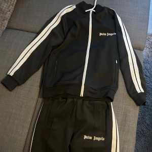Svart träningsjacka från Palm Angels - Säljer en snygg svart dress från Palm Angels med vita ränder längs ärmarna och dragkedja framtill. Går att köpa byxor och tröjan separat. 700 per del för separat köp. Storlek S på båda. 