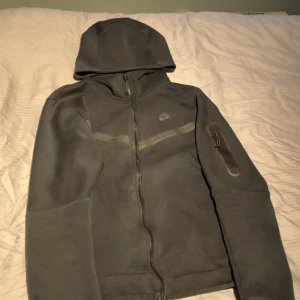 Nike tech svart - Säljer en svart hoodie från Nike i mycket bra skick. Den har en dragkedja framtill och en praktisk ficka med dragkedja på ärmen. Perfekt för höst och vår, och den är superbekväm att ha på sig. Passar både till träning och vardagsbruk.