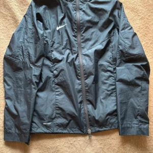 Svart vindjacka från Nike - Säljer en svart vindjacka från Nike i storlek xs. Jackan har en dragkedja framtill och är perfekt för blåsiga dagar. Den är i mycket bra skick och passar bra för träning eller vardagsbruk. Nike-loggan syns tydligt på bröstet. Perfekt för dig som vill ha en stilren och funktionell jacka! 🖤