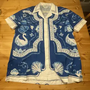 Säljer en supercool blå och vit skjorta från Casablanca med ett unikt mönster av svanar och blommor. Skjortan har korta ärmar och en klassisk krage. Perfekt för sommardagar eller en stilren festlook. Materialet känns lyxigt och bekvämt. Passar perfekt för dig som vill sticka ut med stil! 🌟