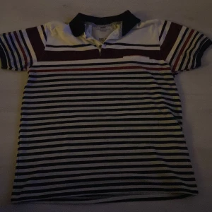 Randig pikétröja från Original Men Fashion - Säljer en snygg randig pikétröja i herrstorlek XL från Original Men Fashion. Tröjan har en klassisk krage och knappar framtill. Färgerna är en mix av vit, svart och vinröd, vilket ger en stilren look. Perfekt för både vardag och fest!