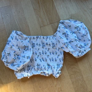 Blommig offshoulder topp från NA-KD - Säljer en superfin blommig offshoulder topp från NA-KD i storlek 34. Den är vit med blå blommor och har en smockad, croppad passform. Perfekt för vardag eller fest! 🌸går att ha off shoulder & som vanligt