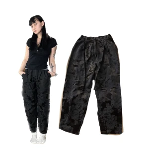 Svarta byxor med blommönster från KappAhl - Snygga svarta byxor från KappAhl med ett elegant blommönster. De är tillverkade i en blandning av viskos och acetat, vilket ger dem en lyxig känsla. Byxorna har en loose passform och är perfekta för både vardag och fest. 