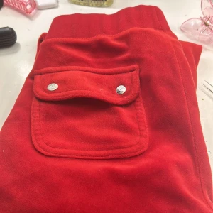 Röda mjukisbyxor från Juicy Couture - Säljer ett par snygga röda mjukisbyxor från Juicy Couture i storlek XXS. Byxorna har en bekväm passform med snörning i midjan och en praktisk ficka med knappar. Perfekta för en avslappnad dag hemma eller en chill dag ute.