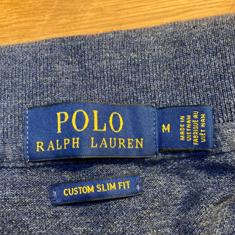 Säljer en snygg grå/blå pikétröja från Ralph Lauren i custom slim fit. Den har en klassisk krage med två knappar och det ikoniska röda broderade logotypen på bröstet. Perfekt för både vardag och lite finare tillfällen. Passar bra till jeans eller chinos!. Tröjor & Koftor.