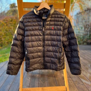 Svart pufferjacka från Polo Ralph Lauren - Säljer en snygg svart pufferjacka från Polo Ralph Lauren i storlek 14-16. Jackan har en quiltad design med dragkedja och två fickor fram. Perfekt för kyliga dagar och har en klassisk stil med det ikoniska röda logotypbroderiet på bröstet. Passar både till vardags och lite finare tillfällen.