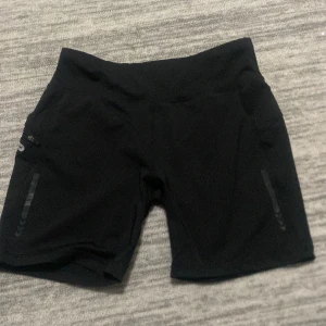 Svarta träningsshorts från New Apparel - Säljer ett par svarta träningsshorts från New Apparel i storlek M. De är tillverkade i ett stretchigt material med dragkedjor på sidorna för extra säkerhet. Perfekta för gymmet eller löprundan. Högmidjade för en bekväm passform.