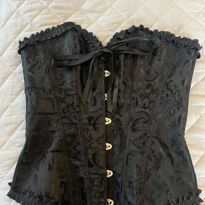 Svart korsett med snörning - Snygg svart korsett med vackert mönster och snörning både fram och bak. Den har volangdetaljer upptill och nedtill som ger en extra touch. Perfekt för festliga tillfällen eller när du vill känna dig extra speciell.