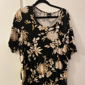 Blommig top från Gina Tricot - Säljer en superfin blommig top från Gina Tricot i storlek L. Den är svart med ett vackert blommönster i beige och gul/guld. Topen har korta ärmar och är perfekt för både vardag och fest. Materialet är mjukt och bekvämt, gjort av viskos och elastan.