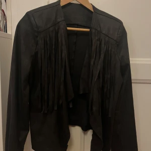 Svart skinnjacka med fransar - Säljer en cool svart skinnjacka med fransar framtill. Perfekt för en edgy look! Jackan har långa ärmar och en öppen design utan knappar eller dragkedja. Passar perfekt till både vardag och fest. Har axelvaddar.