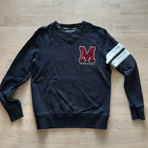 Svart sweatshirt från Morris  - Säljer en mörkblå tröja från Morris R. med ett stort rött 'M' broderat på bröstet. Tröjan har långa ärmar med vita ränder på ena ärmen. Storlek xs