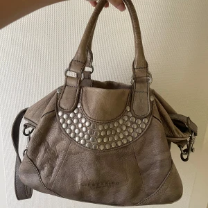 Väska med nitar - Rymlig skinnväska från Liebeskind. Den är mer åt det gröna hållet än vad bilden visar! Köpt second hand och i väldigt använt skick. Innertyget har fläckar men inget man tänker på. Dator får plats! Väskan har både handtag och en avtagbar axelrem. 