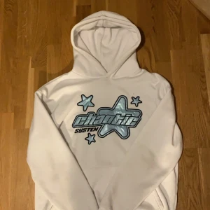 Vit hoodie - Säljer en vit hoodie  med tryck av stjärnor och texten 'chaotic system' i blått och svart. Hoodien har en avslappnad passform och är perfekt för en casual look.
