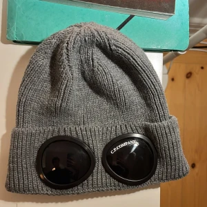 Grå stickad mössa från C.P. Company - Snygg grå stickad mössa från C.P. Company med unika glasögondetaljer framtill. Perfekt för att hålla sig varm och stilren under kyliga dagar. Osäker på äktheten därav det låga priset Hörs av dig vid frågor🙏