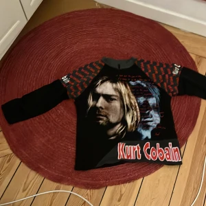 Svart långärmad tröja med Kurt Cobain-tryck - Cool svart tröja med lång ärm och tryck av Kurt Cobain på framsidan. Ärmarna har ett rött och svart mönster med text. Perfekt för fans av grunge och rockmusik.