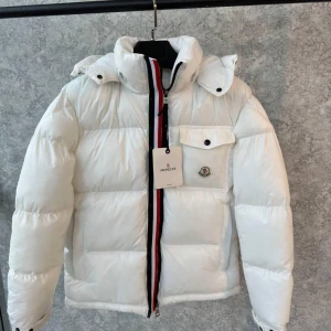 Vit dunjacka från Moncler - Snygg vit dunjacka från Moncler med hög krage och dragkedja i rött, vitt och blått. Jackan har en bröstficka med knapp och Moncler-logga. Perfekt för kyliga dagar.