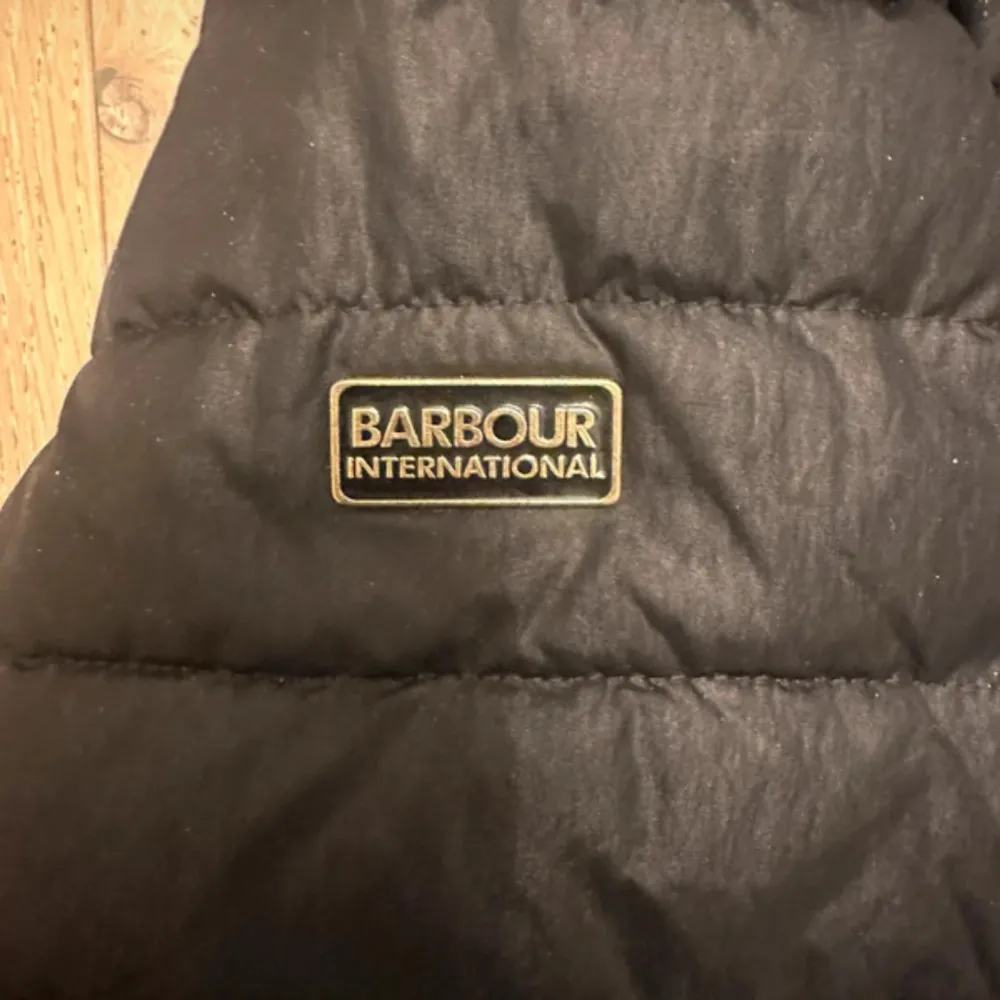 Snygg svart dunjacka från Barbour med pälskantad huva och dragkedja. Jackan har flera praktiska fickor med knappar och ett klassiskt rutigt foder. Perfekt för kyliga dagar.. Takit.