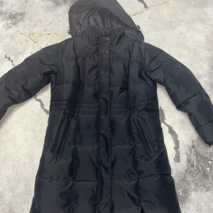 Svart dunjacka från Take Off - Snygg svart dunjacka från Take Off med huva och dragkedja. Perfekt för kalla dagar med sin långa design och praktiska fickor. Jackan har en stilren look och är idealisk för vinter och höst.