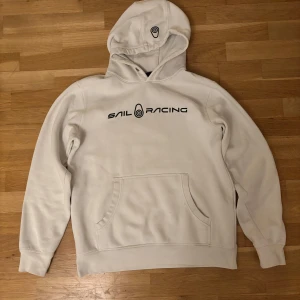 Vit hoodie från Sail Racing - Säljer en stilren vit hoodie från Sail Racing med svart logotyp på bröstet. Den har en klassisk känguruficka och en bekväm huva med dragsko. Perfekt för en avslappnad stil. Saknar snören