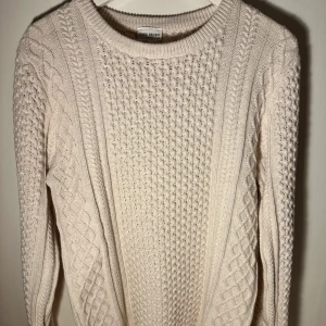 Beige stickad tröja från john henric - Mysig beige stickad tröja från john henric med ett klassiskt flätmönster. Perfekt för kyliga dagar med sin varma och bekväma design. Tröjan har långa ärmar och rund halsringning.