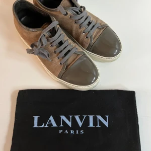 Bruna sneakers från Lanvin - Snygga bruna sneakers från Lanvin med grå snörning och detaljer i lack. Skorna har en vit sula och är tillverkade i mocka med en glansig tåhätta. Perfekta för en stilren look.