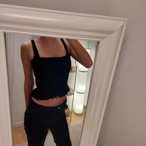 Svart croppad topp från H&M - Säljer en svart croppad topp från H&M med breda axelband och volangdetalj vid nederkanten. Toppen har en smockad design som ger en tight passform. Perfekt för en stilren look.