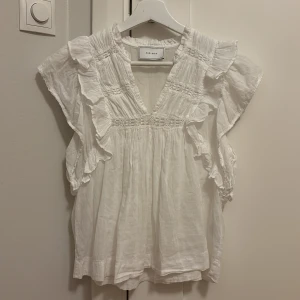 Vit blus från Neo Noir - Charmig vit blus från Neo Noir med volangdetaljer och v-ringning. Blusen har korta ärmar och en luftig passform, perfekt för varmare dagar. Blusen är använd fåtal gånger därför i väldigt fint skick!