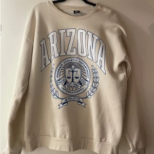 Beige sweatshirt med Arizona-tryck - Säljer en beige sweatshirt med stort Arizona-tryck i blått på framsidan. Tröjan har långa ärmar och en rund halsringning. Perfekt för en avslappnad stil.
