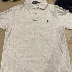 Grå pikétröja från Ralph Lauren - Säljer en stilren grå pikétröja från Ralph Lauren med broderad logga på bröstet. Tröjan har korta ärmar och en klassisk krage med knappar. Perfekt för en avslappnad men ändå snygg look.