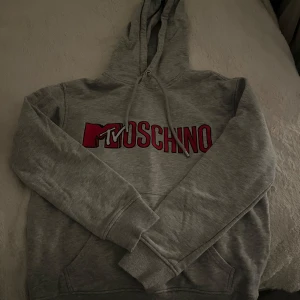 Grå hoodie från Moschino - Säljer en grå hoodie från Moschino med röd och svart logga på framsidan. Tröjan har en klassisk huva med dragsko och ribbade muddar vid ärmslut och nederkant. Perfekt för en avslappnad stil. Super snygg!! St S,  passar mig som har s-m. Knappt använd alltså som ny
