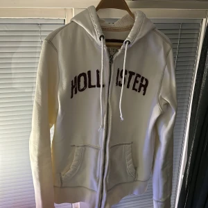 Vit hoodie från Hollister - Säljer en vit/cream färg hoodie från Hollister med dragkedja och har en justerbar huva och fickor framtill. Perfekt för en avslappnad stil. Tröjan har en sliten look i bra kvalité. Hör av dig om du har frågor:)