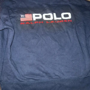 Mörkblå sweatshirt från Ralph Lauren - Säljer en snygg mörkblå sweatshirt från Ralph Lauren med det klassiska POLO-trycket i vitt och rött på framsidan. Perfekt för en avslappnad stil. Tröjan har rund hals och långa ärmar.