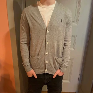 Grå kofta från Ralph Lauren - Snygg grå kofta från Ralph Lauren med knappar framtill och två fickor. Den har en klassisk design med ribbade muddar och en liten broderad logotyp på bröstet. Perfekt för lager-på-lager-stil.