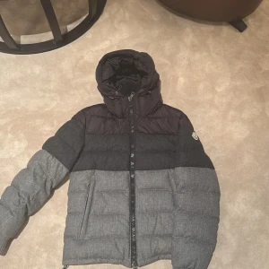 Grå och svart dunjacka från Moncler - Säljer en snygg grå och svart dunjacka från Moncler. Jackan har en quiltad design med dragkedja och ribbade muddar vid ärmsluten. Perfekt för kyliga dagar med sin varma och stilrena look.. Litet slitage längst ner på ärmen. 