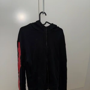 Svart hoodie med röda detaljer - Snygg svart hoodie med röda och vita detaljer på ärmarna och huvan. Perfekt för en casual look med dragkedja framtill och långärmad design.