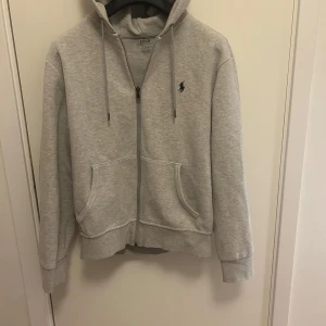 Grå hoodie från Polo Ralph Lauren - Säljer en stilren grå hoodie från Polo Ralph Lauren med dragkedja och det klassiska logotypbroderiet på bröstet. Tröjan har en bekväm passform med långa ärmar och en praktisk huva med dragsko.
