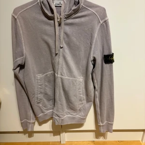 Grå/lila huvtröja från Stone Island - Säljer en stilren grå/lila huvtröja från Stone Island med dragkedja och fickor framtill. Tröjan har en ikonisk patch på ärmen och är perfekt för en avslappnad look.