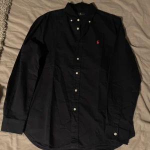 Svart skjorta Ralph Lauren - En svart Ralph lauren skjorta med röd logotyp som är i nyskick. Säljes då den ej används. Knapp fattas ej, bara jag som glömde knäppa sista. Köptes på zalando för 1695kr, kom med bud om mitt pris inte passar ☺️