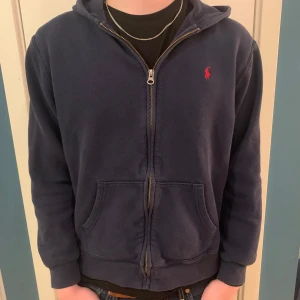 Ralph Lauren full zip - Säljer en snygg mörkblå hoodie från Ralph Lauren med dragkedja och röd logga på bröstet. Tröjan har en klassisk design med huva och fickor framtill. Perfekt för en avslappnad stil. Storlek XL barn (motsvarar xs‐s)  nypris: 990kr