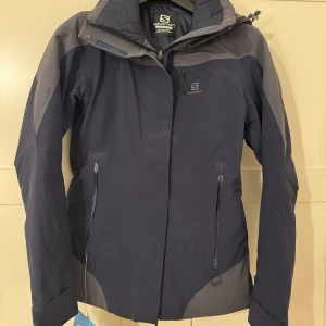 Skidjacka dammodell NY! Salomon - Oanvänd (lappar kvar) snygg skidjacka lätt figursydd. Dammodell. Storlek XS. Grå och gråblå. Avtagbar luva. Märke Salomon. Fodrad. Nypris 3600kr.
