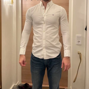 Vit skjorta från GANT - Säljer en stilren vit skjorta från GANT med broderad logotyp på bröstet. Skjortan har en klassisk krage och knappar framtill. Perfekt för en elegant look.