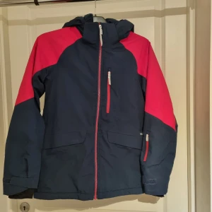 Skidjacka från Didriksons stl.160 - Säljer en mörkblå och cerise skidjacka / vinterjacka från Didriksons. Jackan är 100% vattentät, fodrad och har en praktisk huva. Den har dragkedja framtill och flera fickor med dragkedjor. Snölås. Perfekt för skidåkning eller utomhusaktiviteter i kyligare väder.